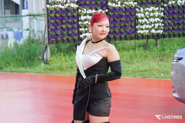 Young woman with bright red hair posing outdoors in a black and white dress against a wall of purple and white flowers.”] ,